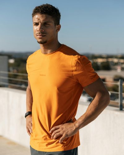 Fit man in orange shirt poses confidently on rooftop in Austin, Texas during a sunny day.